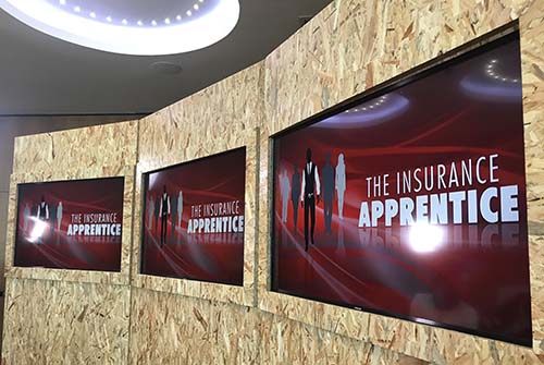 A wall with 3 large screen displaying "The Insurance Apprentice" title.